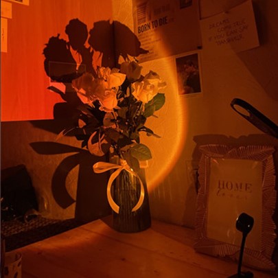 Bouquet de roses blanches, dans un vase transparent avec un ruban nœud blanc également, posé sur un bureau à côté d'un cadre photo, éclairé par  la lampe  coucher de soleil LED USB portable. On voit accroché au mur une photo, un papier avec une phrase écrite et un article de journal en anglais.