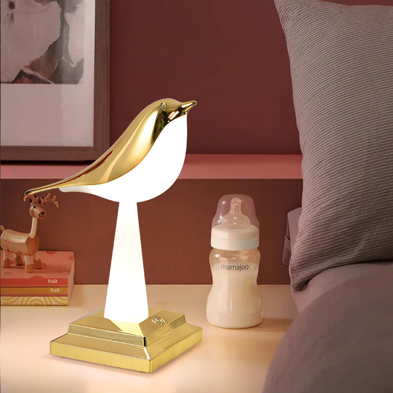 Une lampe en forme d'oiseau dorée et blanche, posée sur une table de chevet à côté d'un lit. Sur la table de chevet il y a aussi un biberon de lait et des livres.